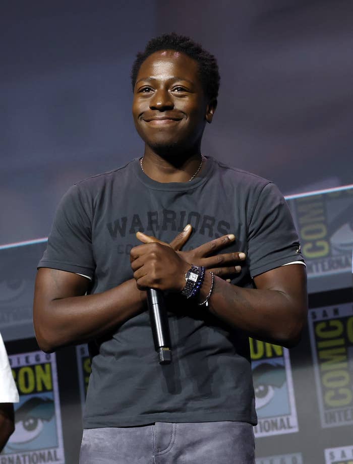 Person smiling on stage at a TV and Movies event, holding a microphone. Wearing casual T-shirt and jeans, standing in front of branded backdrop
