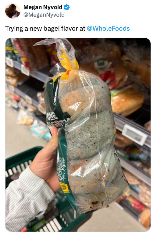 Seseorang memegang sebungkus bagel dengan tampilan berjamur, diberi label rasa baru, di lorong toko kelontong