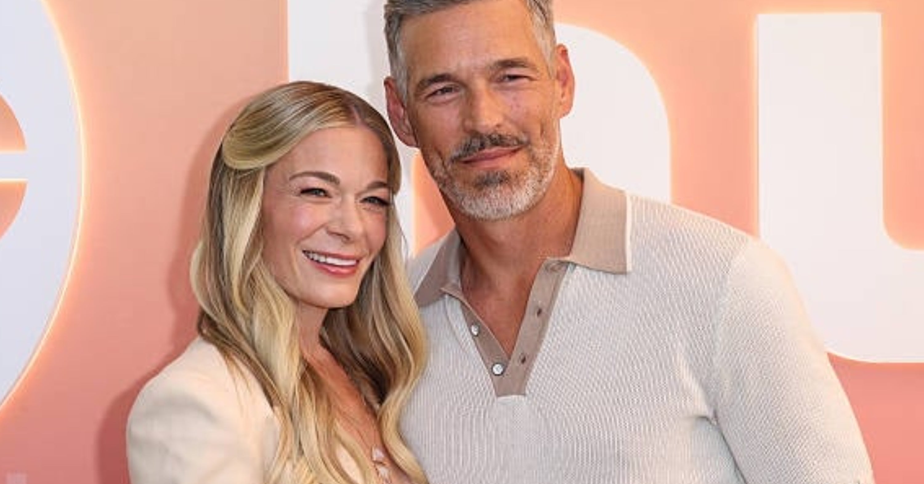 Two people smiling at a media event. The woman is wearing a light-colored top and the man is in a light polo shirt