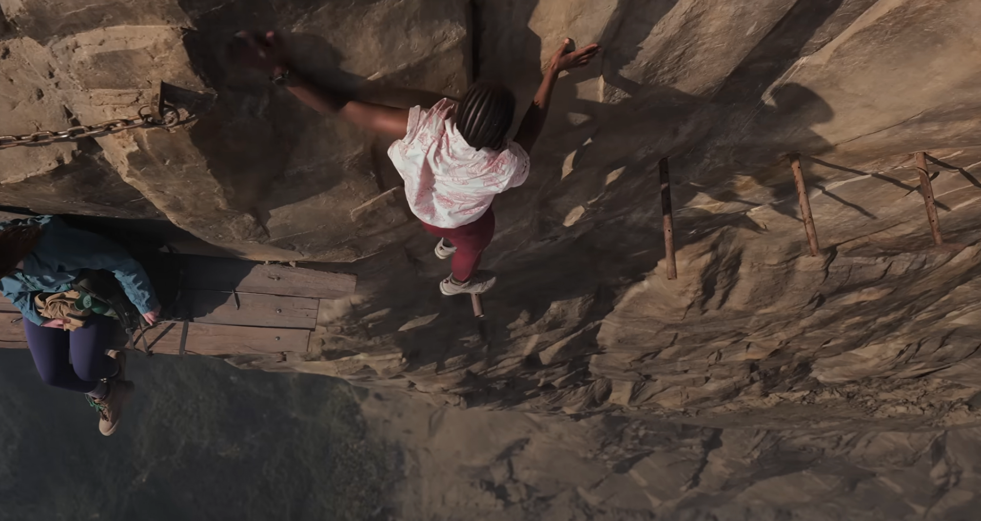 A person sitting on a wooden plank, another balancing on metal beams sticking out of a cliff in "Fall 2: Deadpoint"