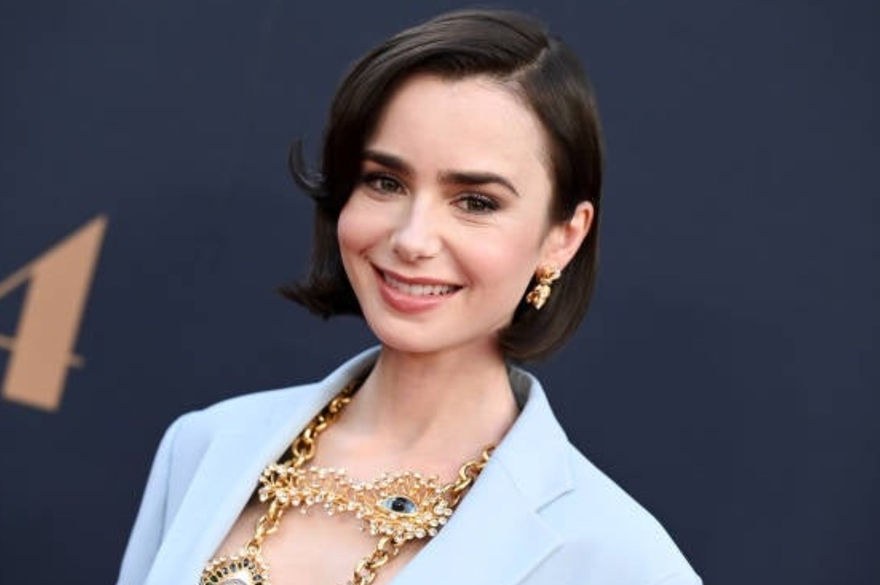 A woman smiles on a red carpet, wearing a light suit and ornate gold jewelry