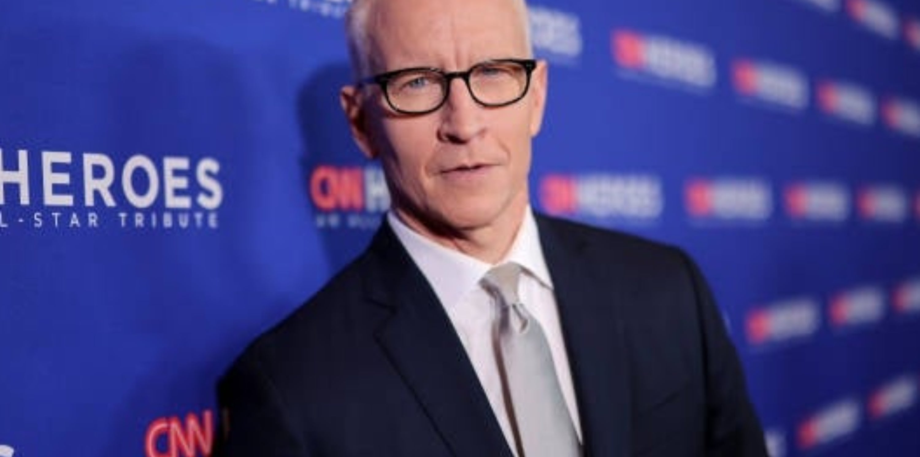 A person in a suit stands on a CNN Heroes event carpet, wearing glasses and a tie, looking directly at the camera
