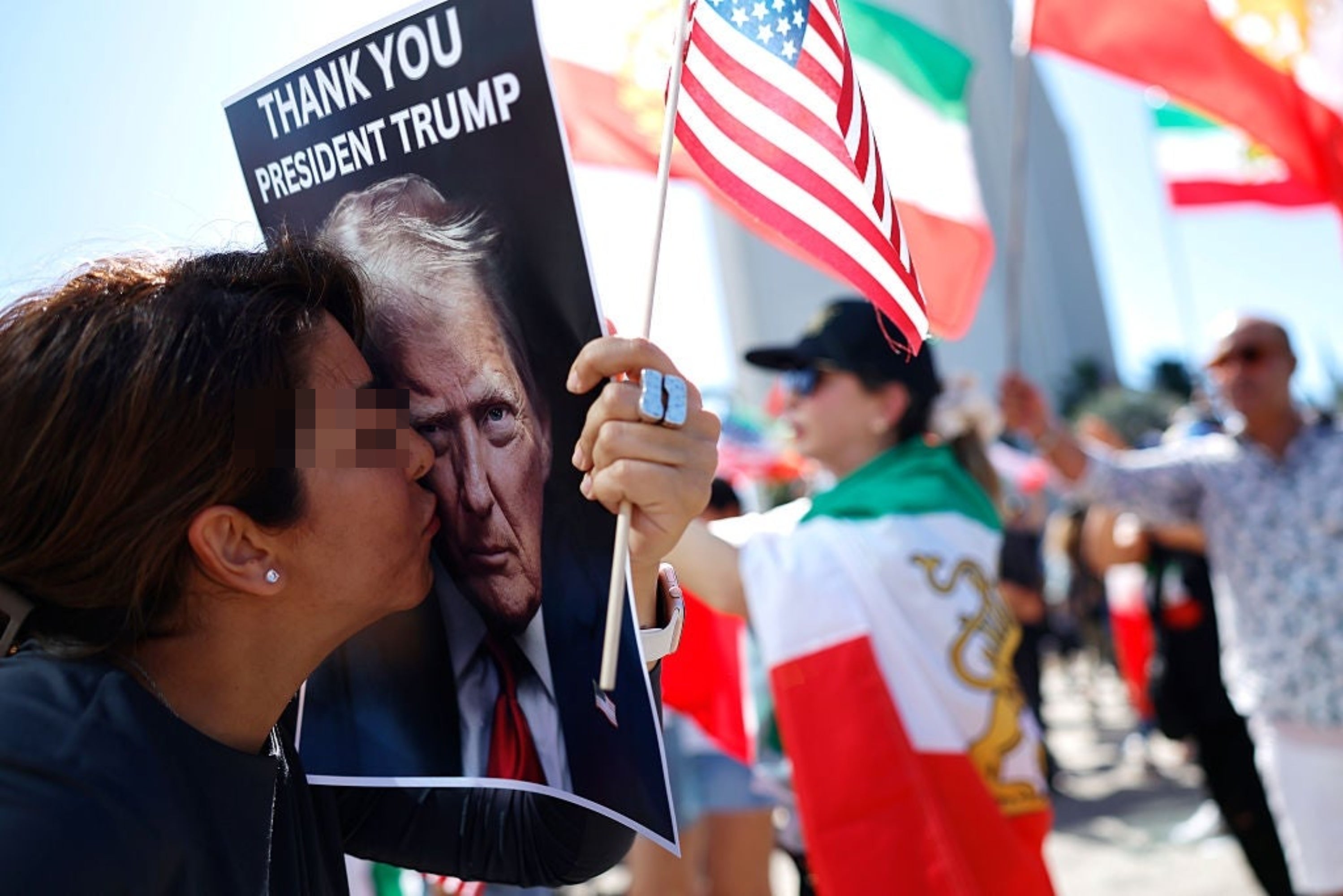 En kvinne kysser en plakat av president Trump på et møte, holder et amerikansk flagg, med folk som vifter med forskjellige nasjonale flagg i bakgrunnen