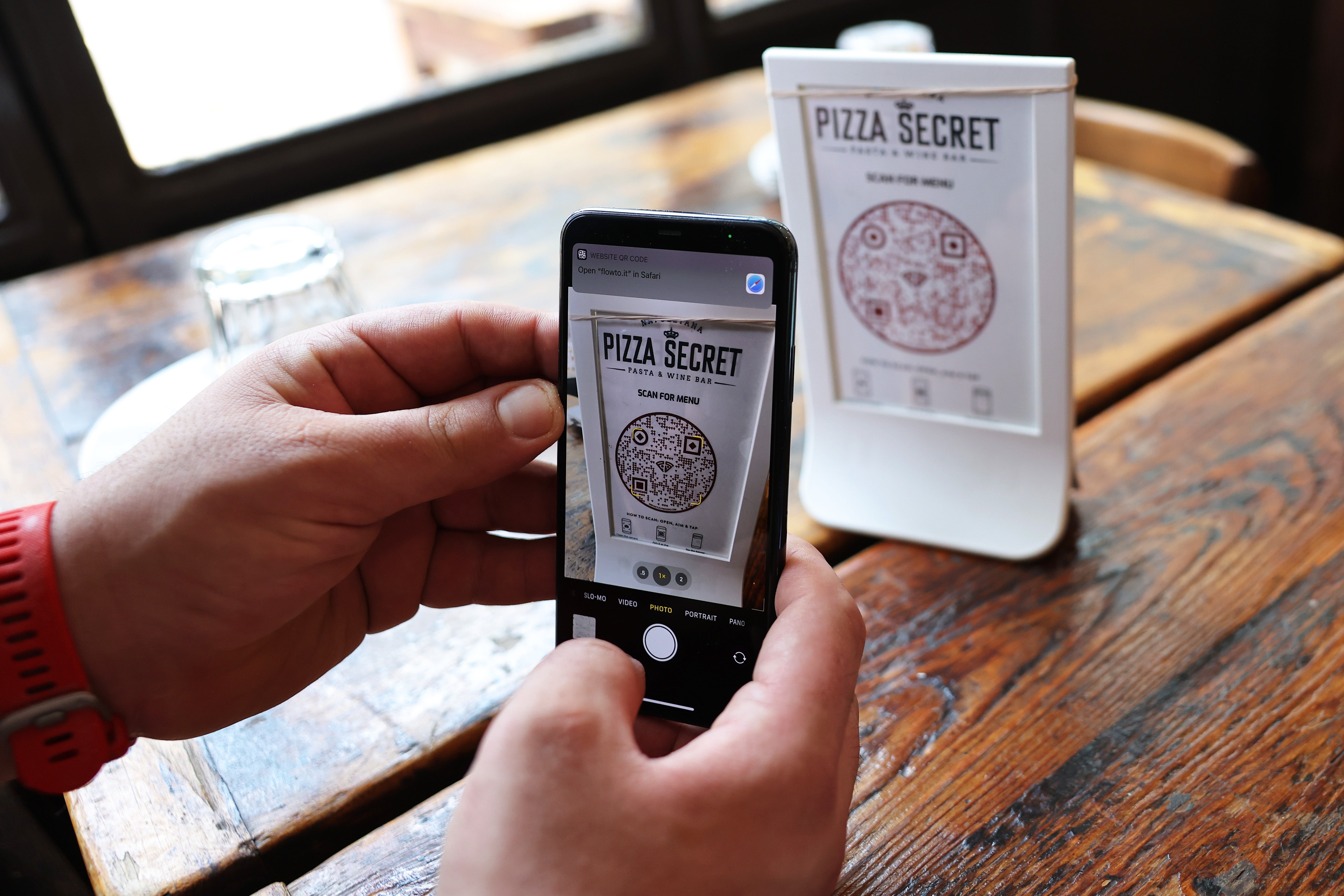 Person som fotograferer en "Pizza Secret" meny med en telefon, med en QR-kode på et trebord