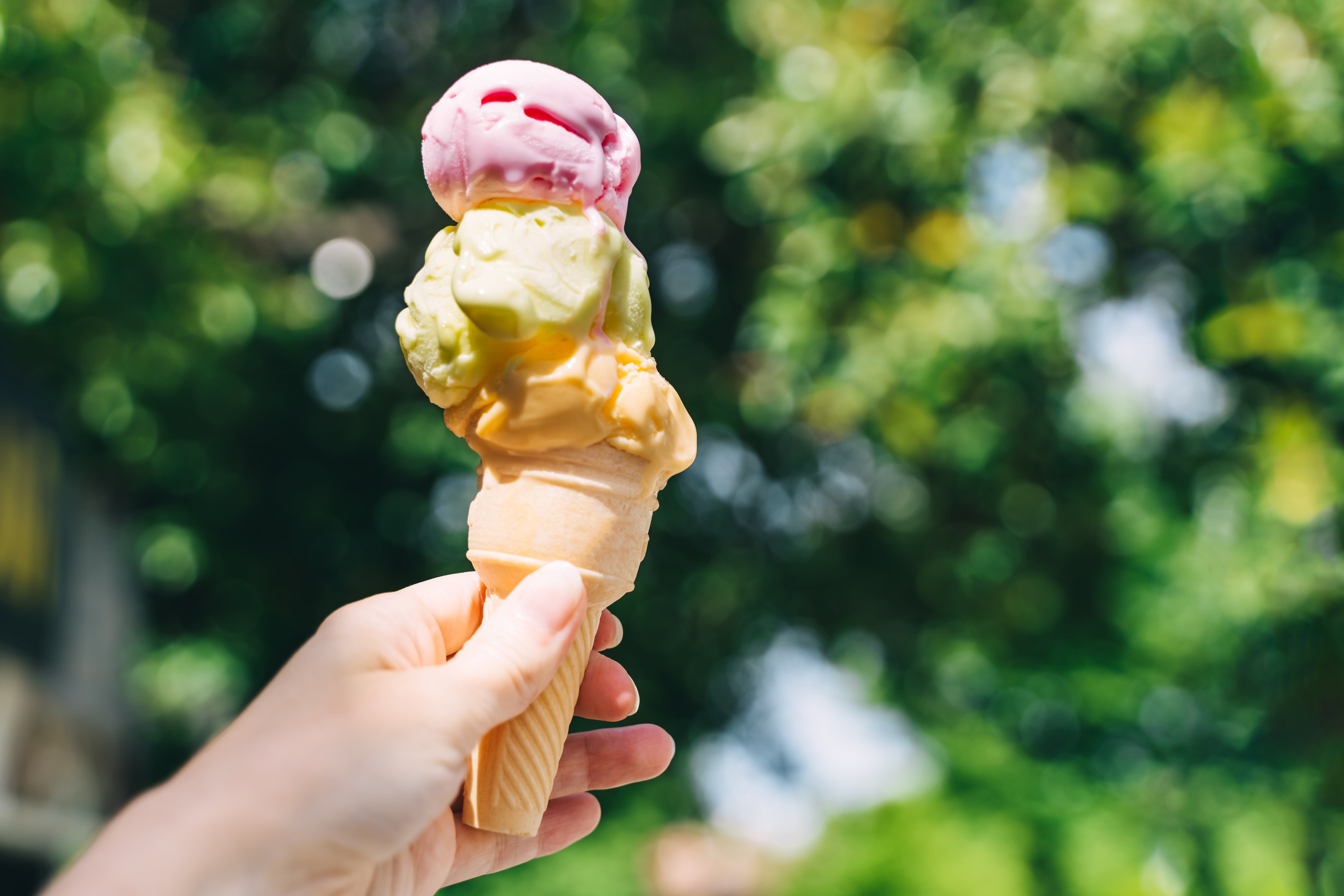 En hånd som holder en smeltende triple-scoop iskrem mot en uskarp utendørs bakgrunn