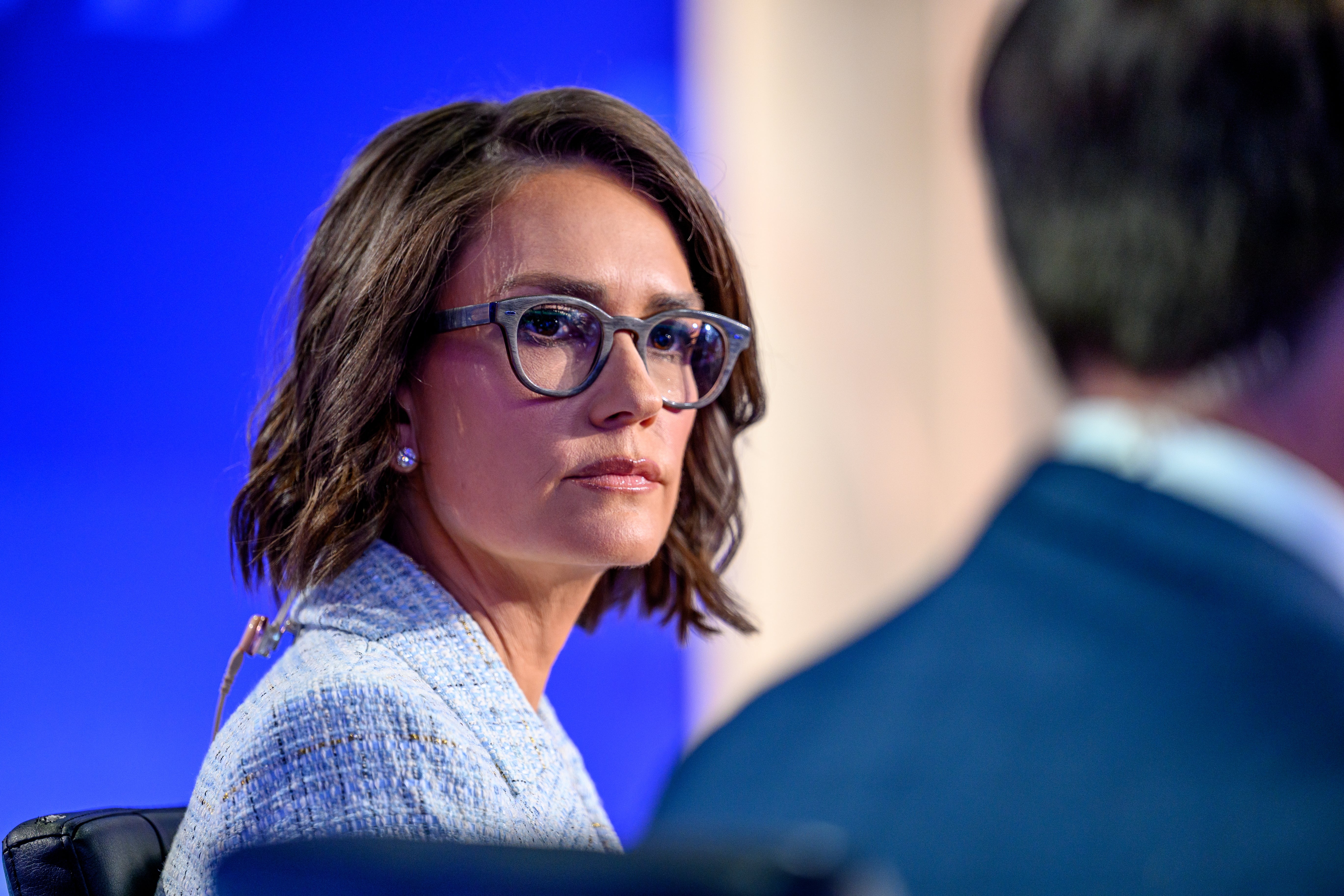 Person med briller i profesjonelle omgivelser, iført en teksturert blazer, ser fokusert ut under en diskusjon eller et panel