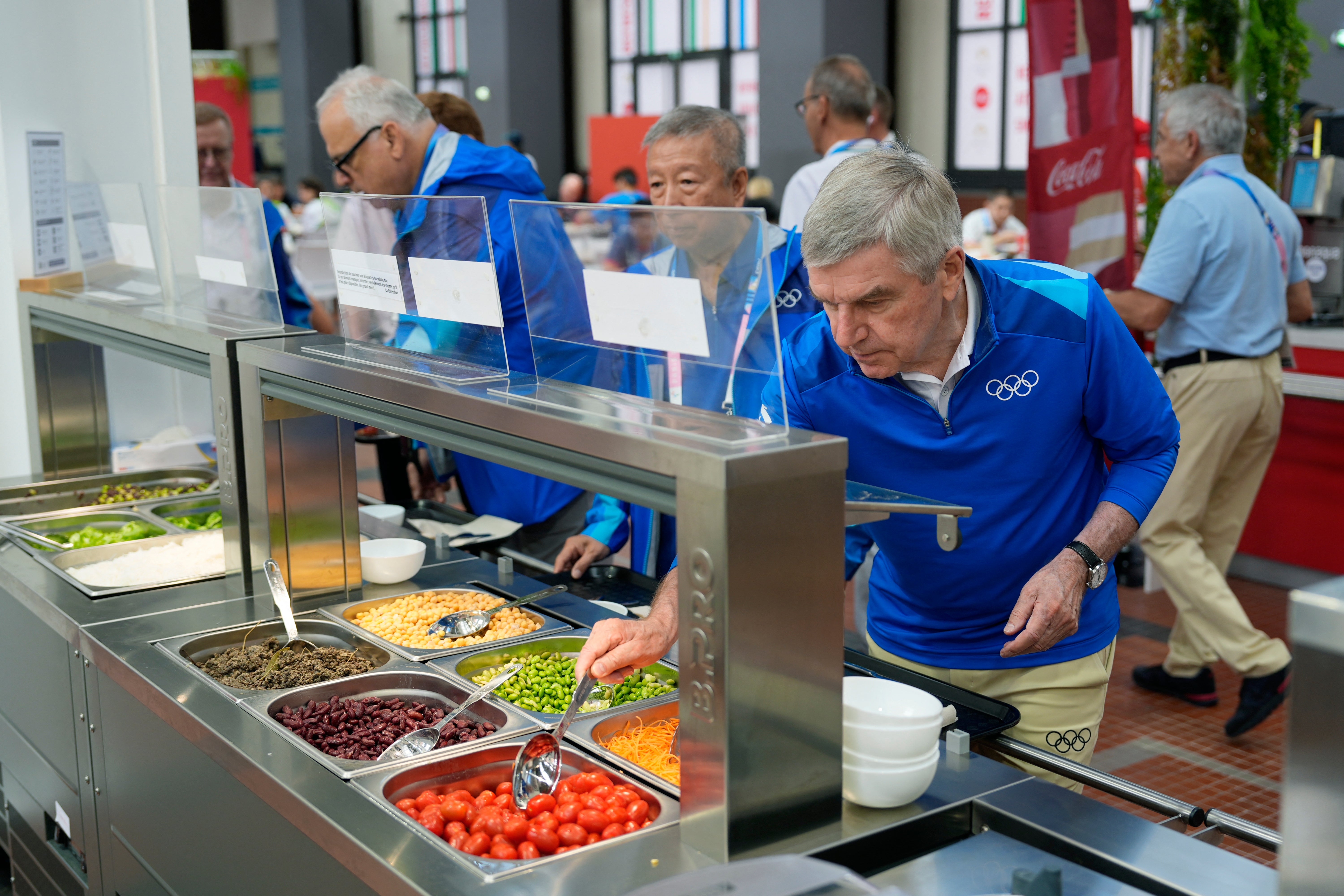 Flere personer i blå OL-skjorte serverer seg selv på en buffet med ulike matvarer i kafeteria-setting
