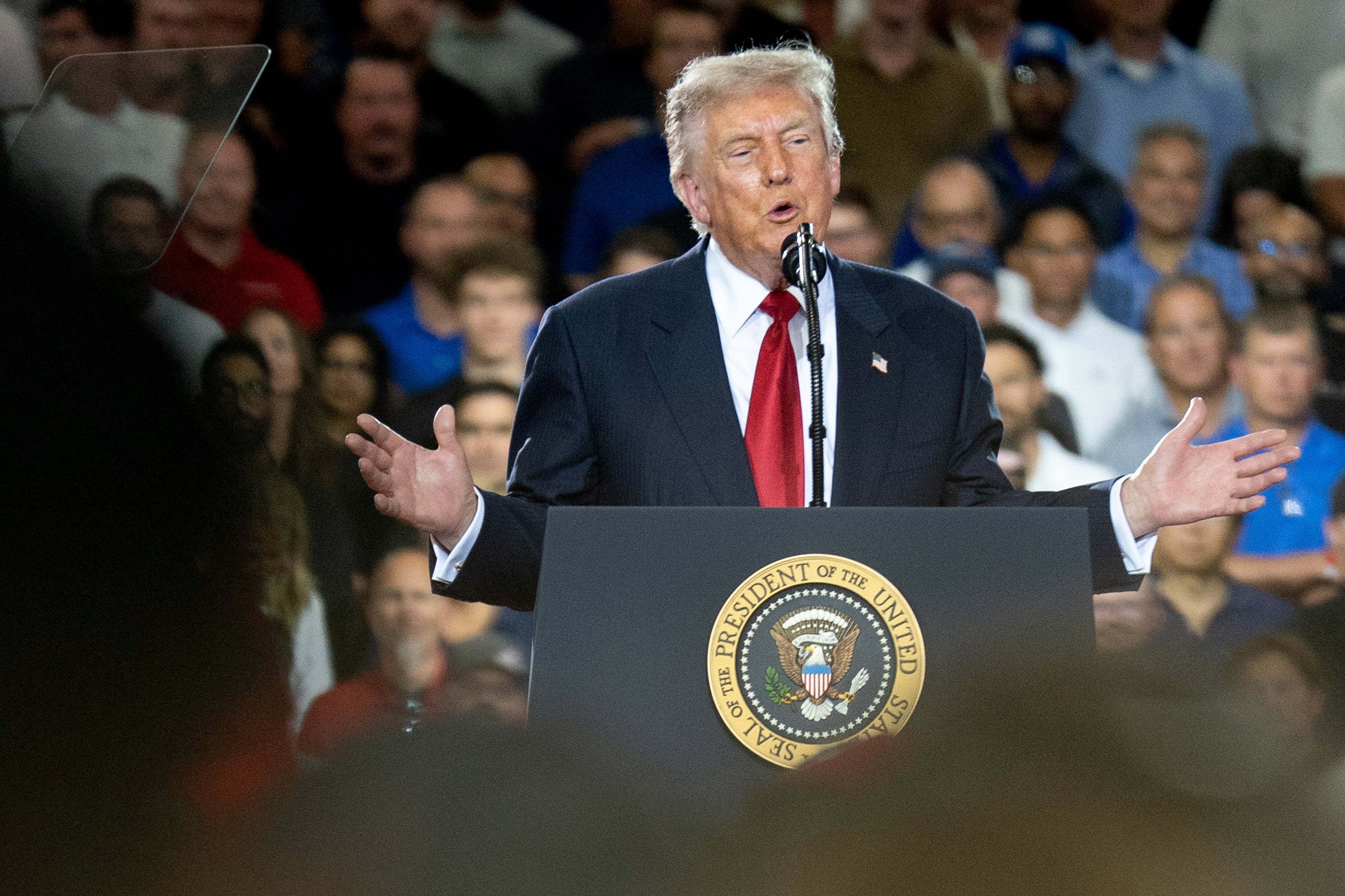 En foredragsholder på et podium med presidentens segl henvender seg til et stort publikum i et overfylt lokale