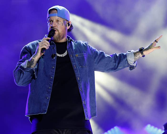 Person on stage in denim jacket and baseball cap, holding a microphone, gesturing with one hand during a performance