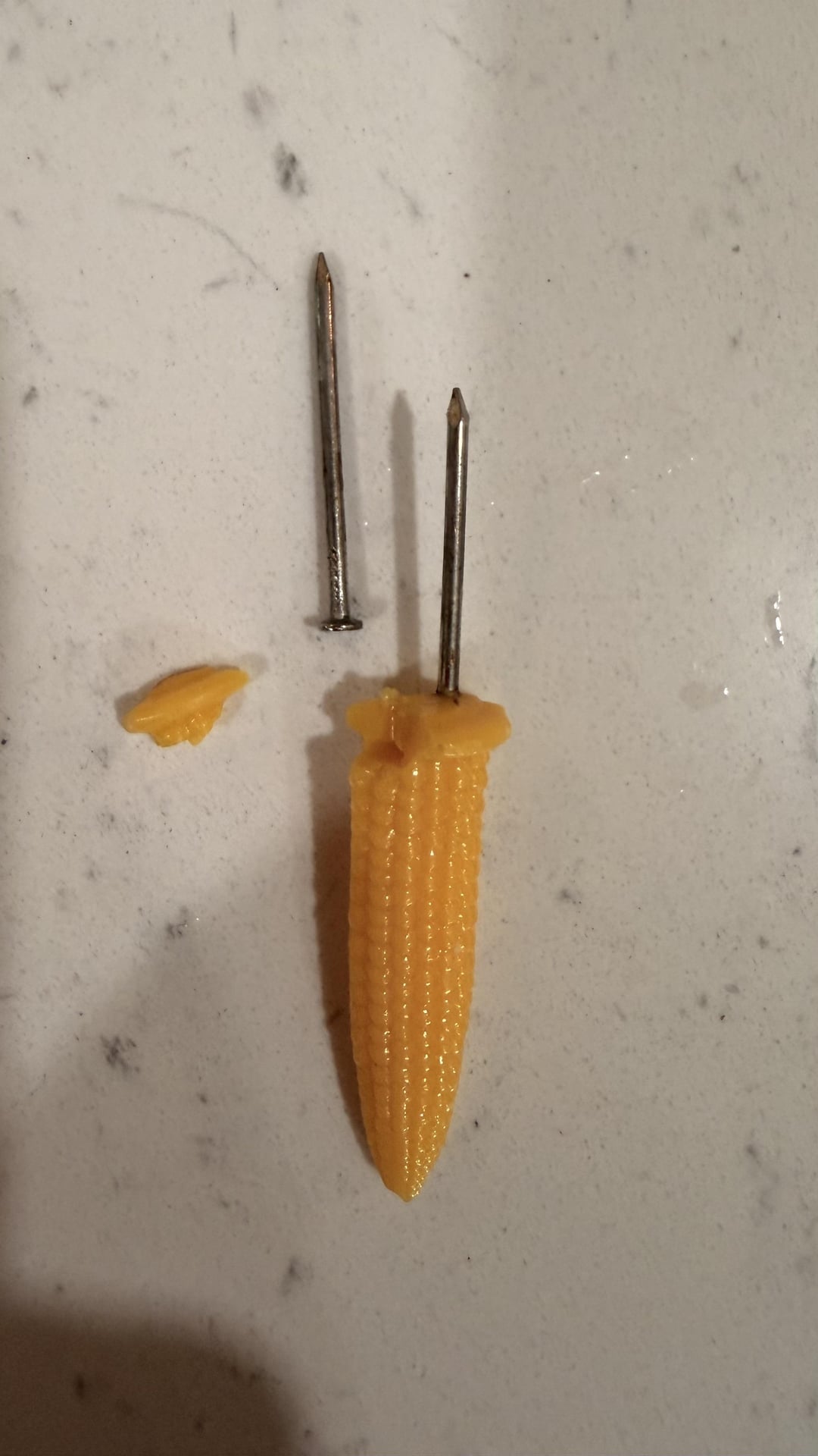 Baby corn pierced by two nails, one nail with only half the length shown. A small piece of corn is cut off beside it