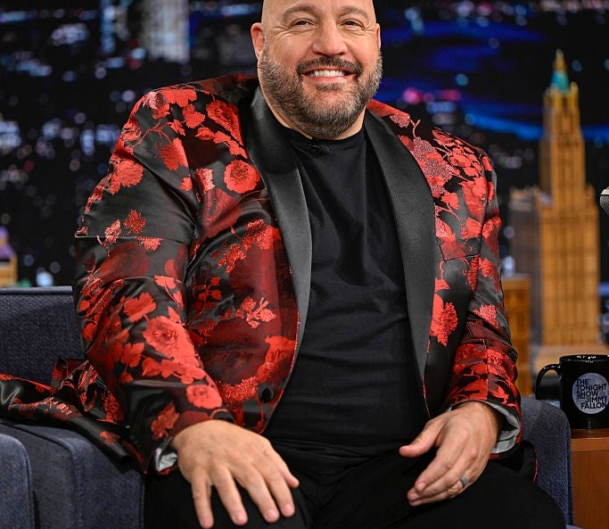 Person sitting on a talk show set, wearing a black shirt and a floral-patterned blazer. Smiling at the camera