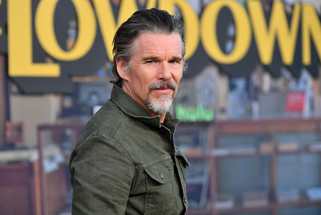 Man in a green jacket with a neatly groomed beard poses on a media event carpet, bold yellow letters partially visible in the background
