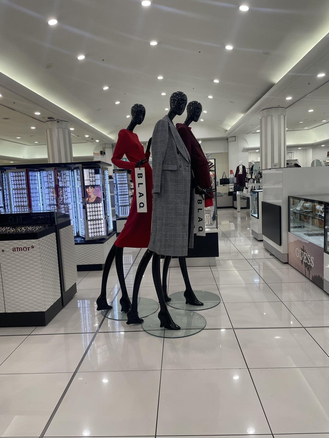Three mannequins in a store display wear red dresses and a gray coat, each holding a shopping bag labeled "Blaq."