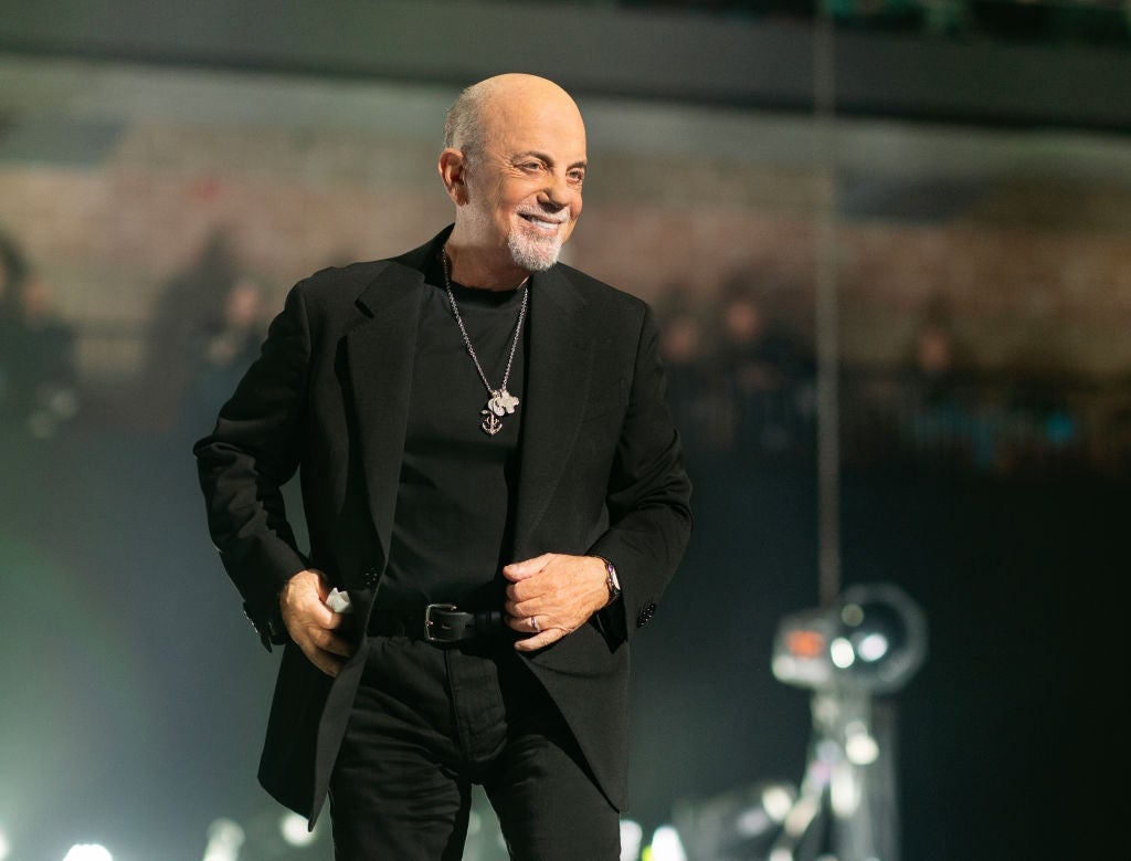 A smiling man on stage, wearing a black suit and turtleneck, accessorized with a long necklace