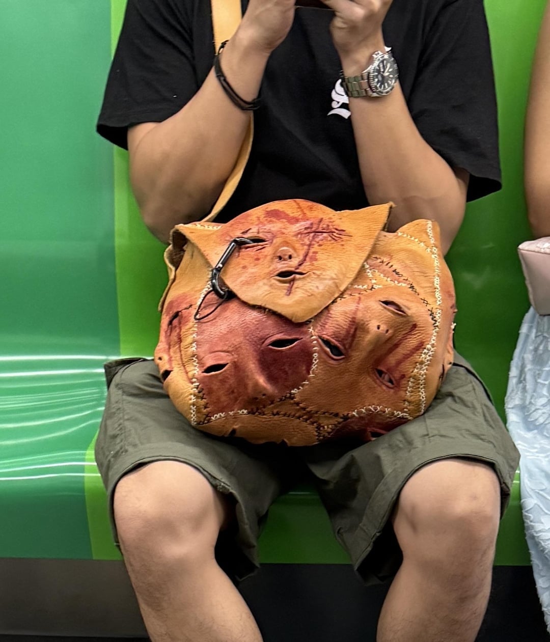 Person sitting on a subway with bag featuring stitched faces design, wearing shorts and a t-shirt, holding a phone