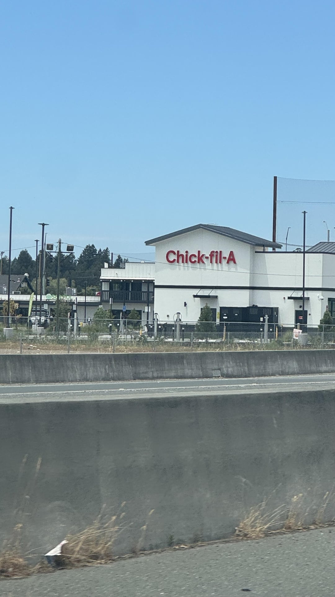 Chick-fil-A restaurant exterior seen from a distance, across a freeway barrier, with clear sky above