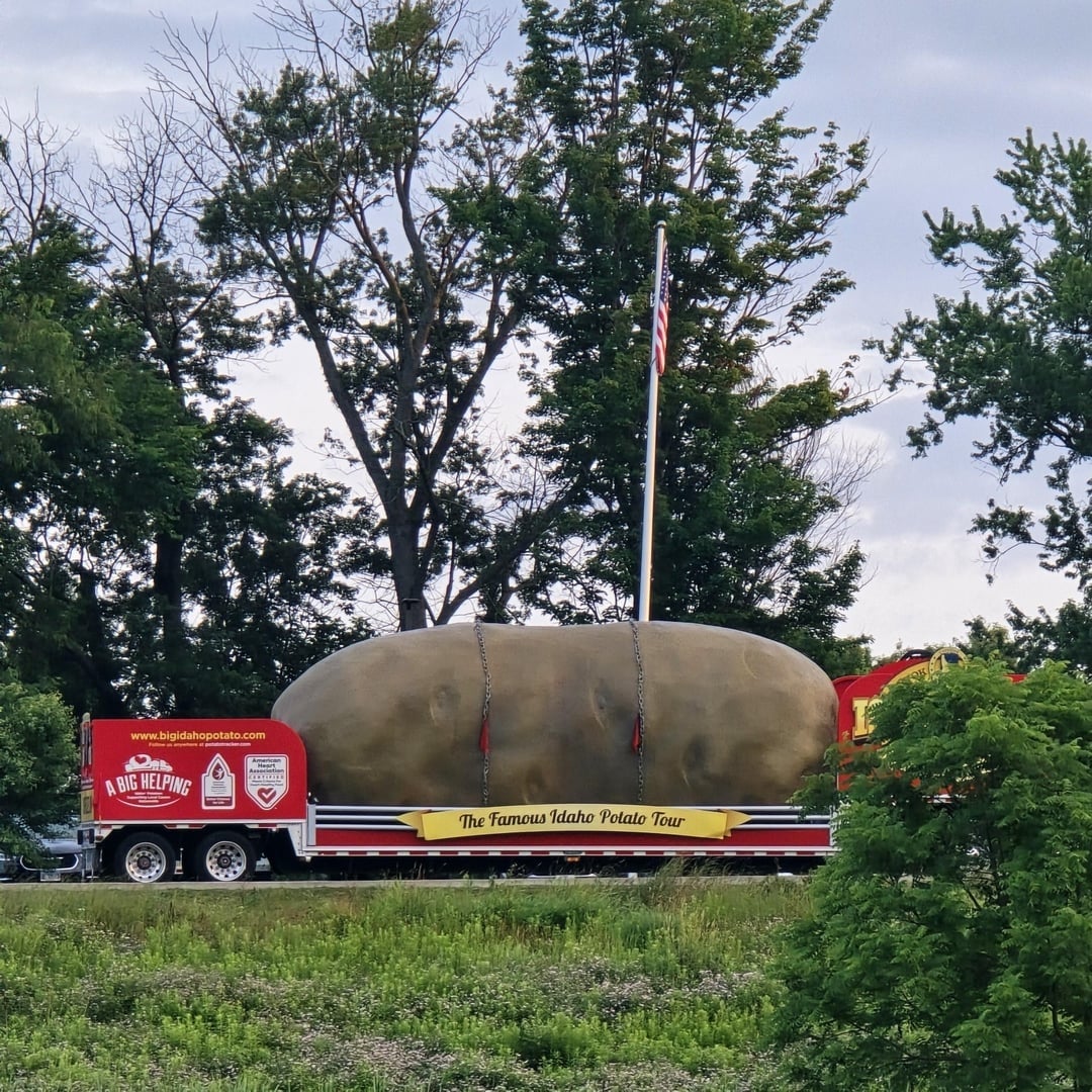Giant potato replica on a flatbed trailer with "The Famous Idaho Potato Tour" sign, parked in a grassy area with trees in the background