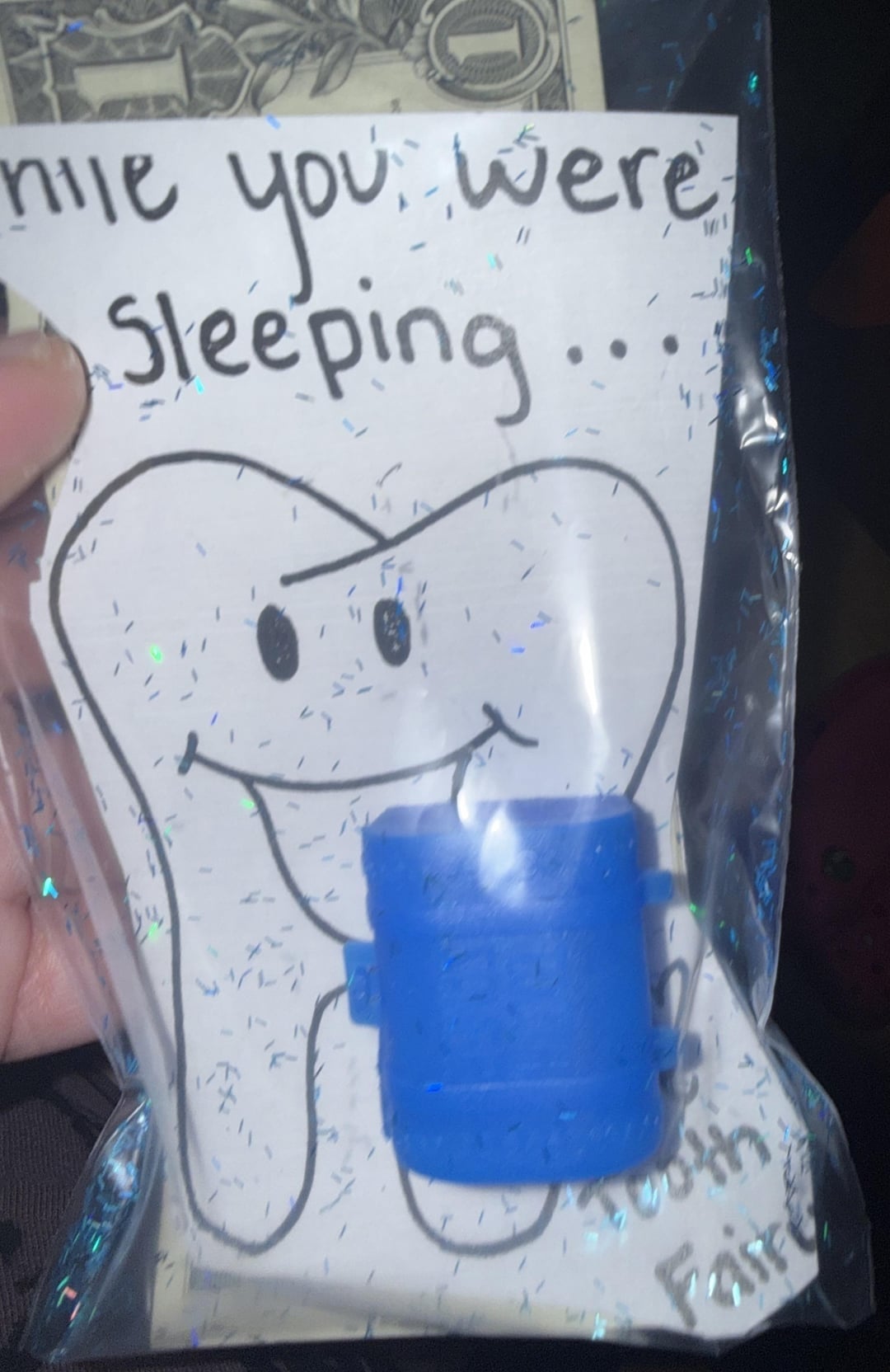 Cartoon tooth with a smile holds a blue mini trash can labeled "Tooth Fairy." Text partially reads "While you were sleeping…" with a dollar bill in background