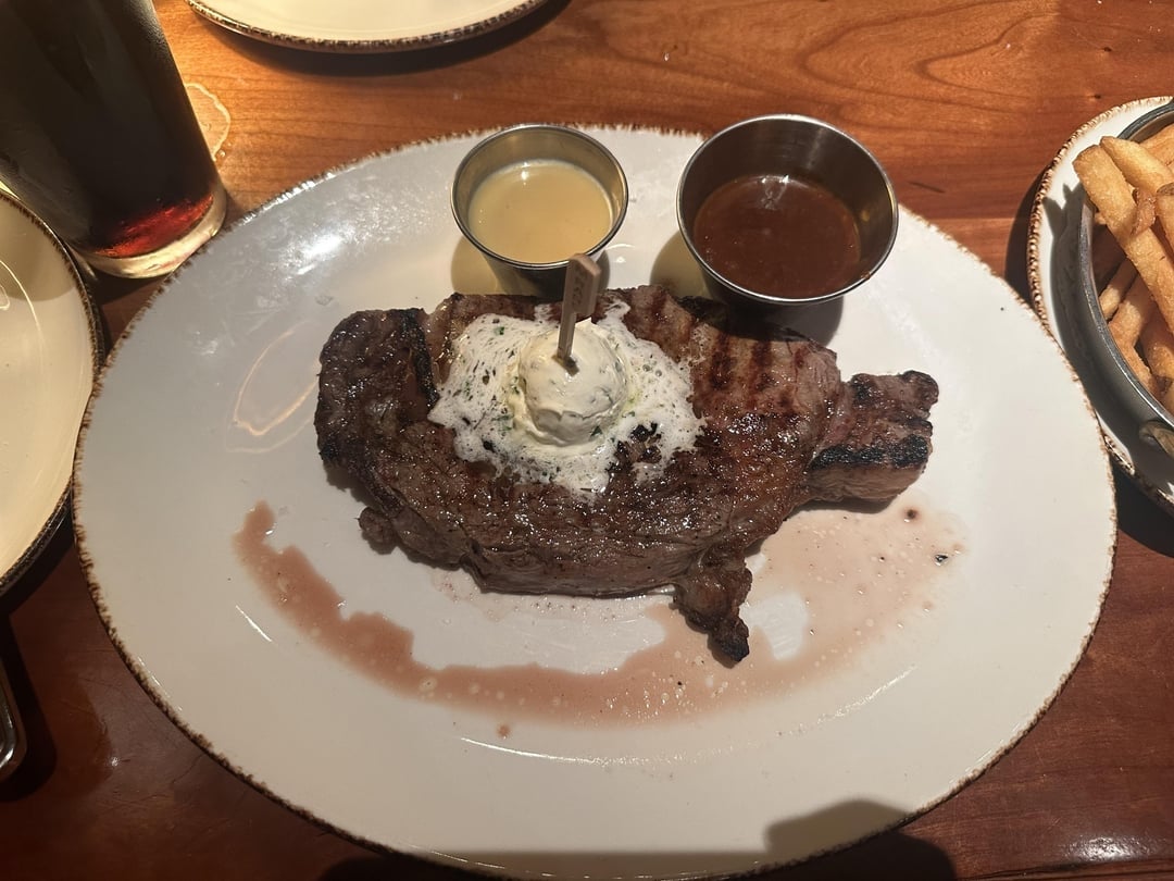 A grilled steak topped with herb butter, served with two sauces, on a white plate. Fries are visible to the side