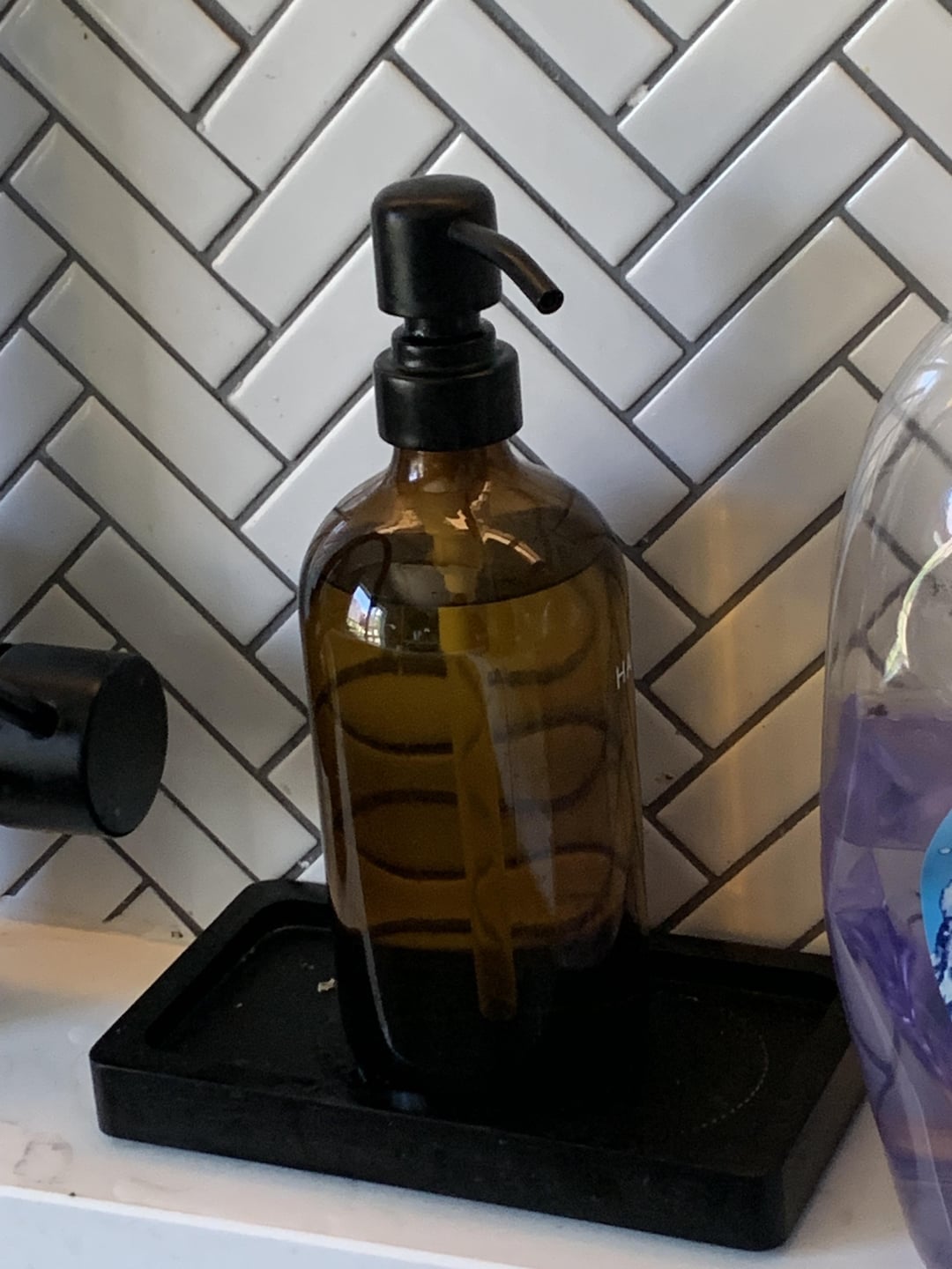 Brown glass soap dispenser with pump on a counter, placed against a white tiled backsplash