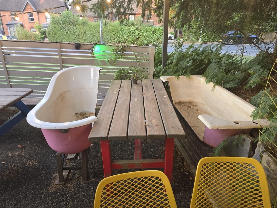 Two bathtubs repurposed as seating around a wooden table in an outdoor setting with greenery in the background