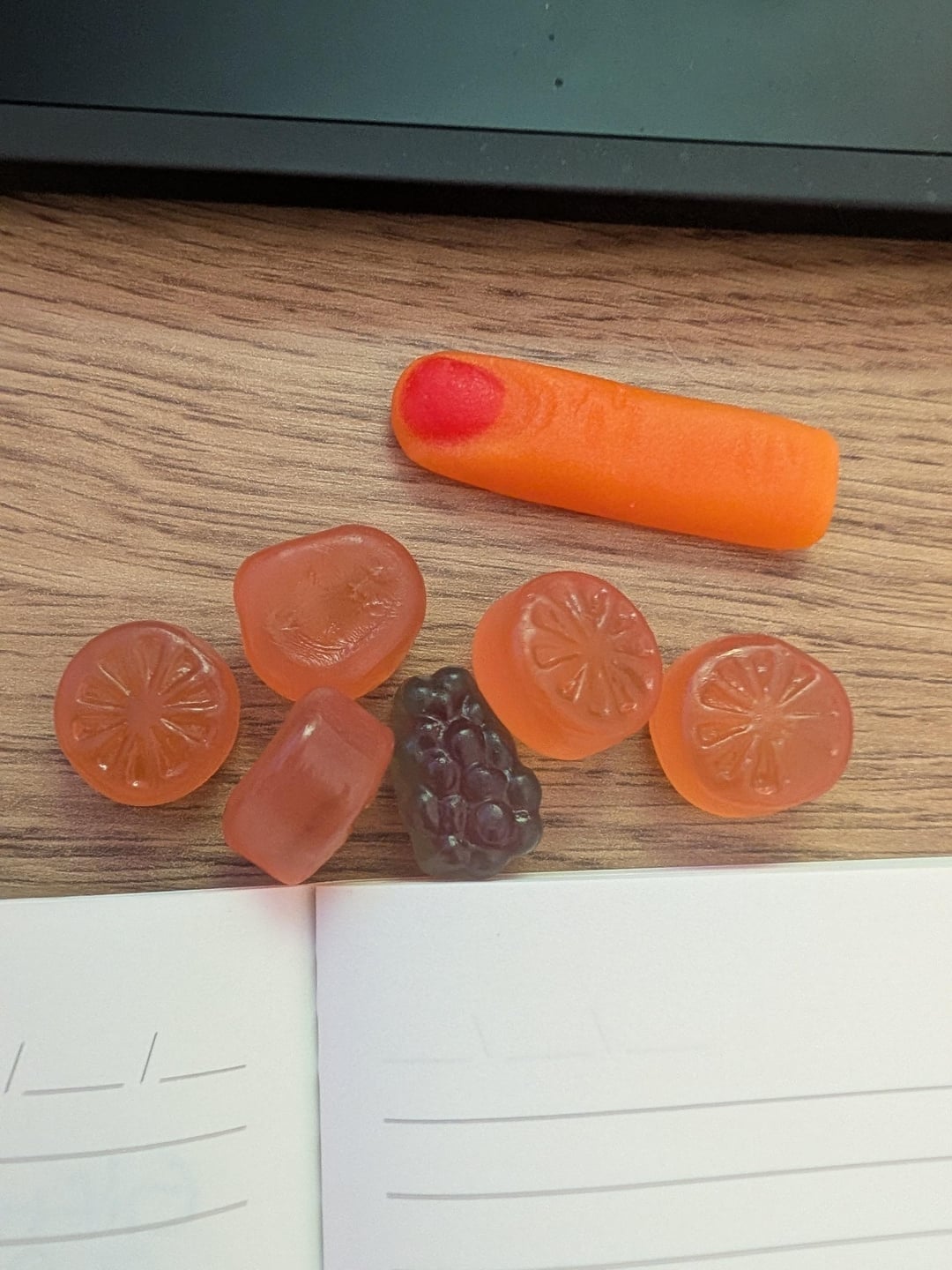 Assorted fruit-flavored gummy candies on a wooden surface, near partially visible notebook pages