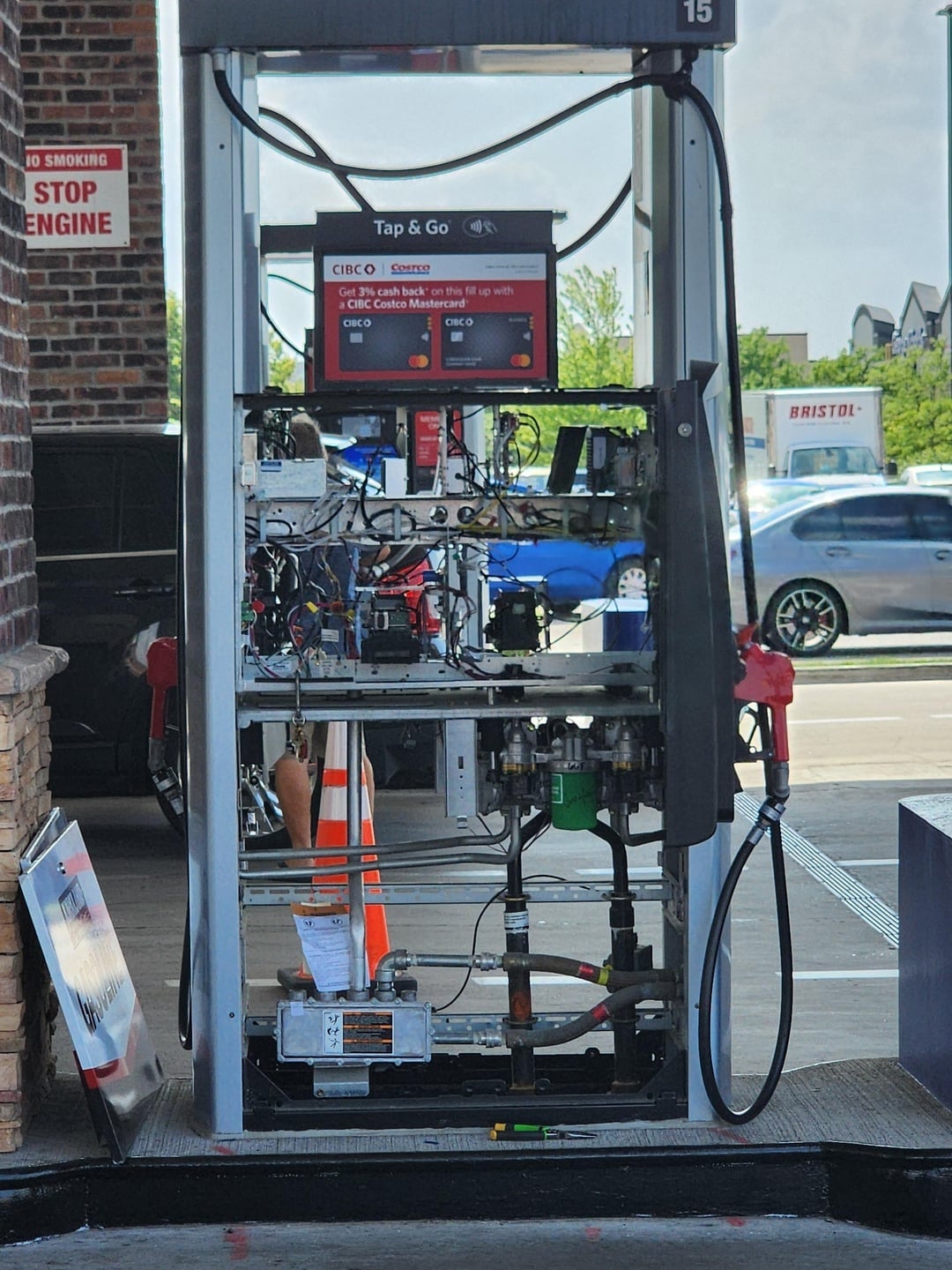 Gas pump with exposed internal components, caution cone nearby. Parts and hoses visible. No people present. Sign reads "Stop engine."