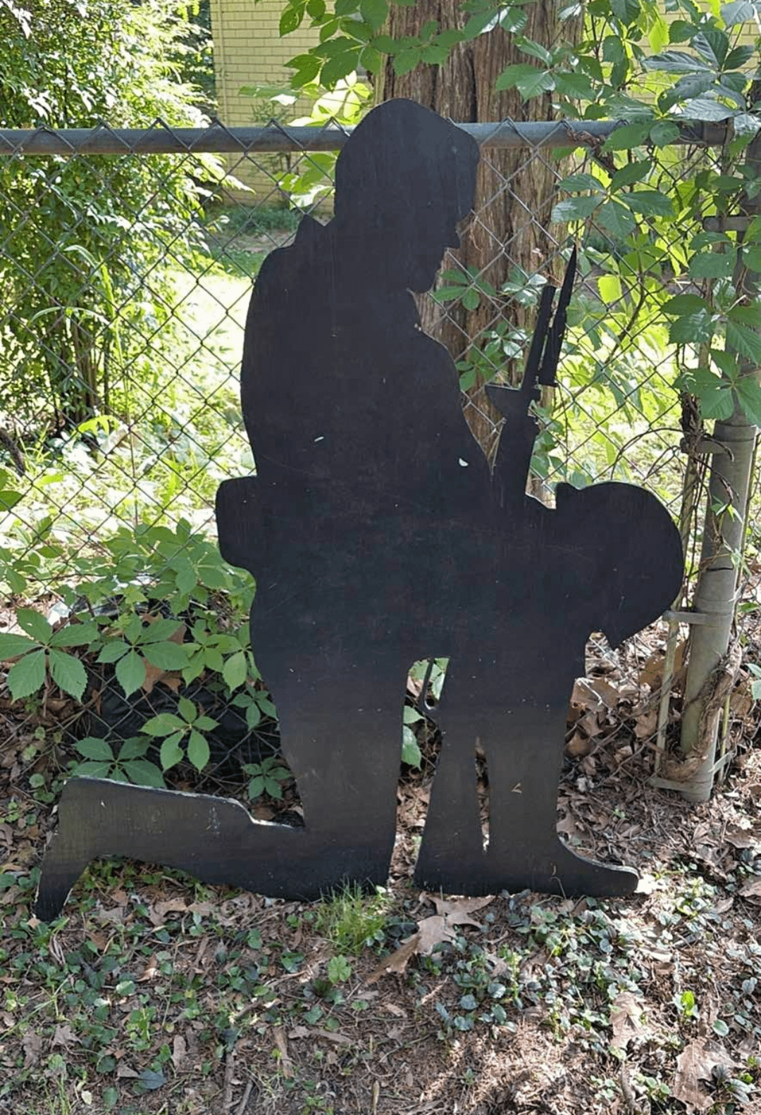 Silhouette of a soldier kneeling with a rifle, set against a garden backdrop with a chain-link fence and greenery