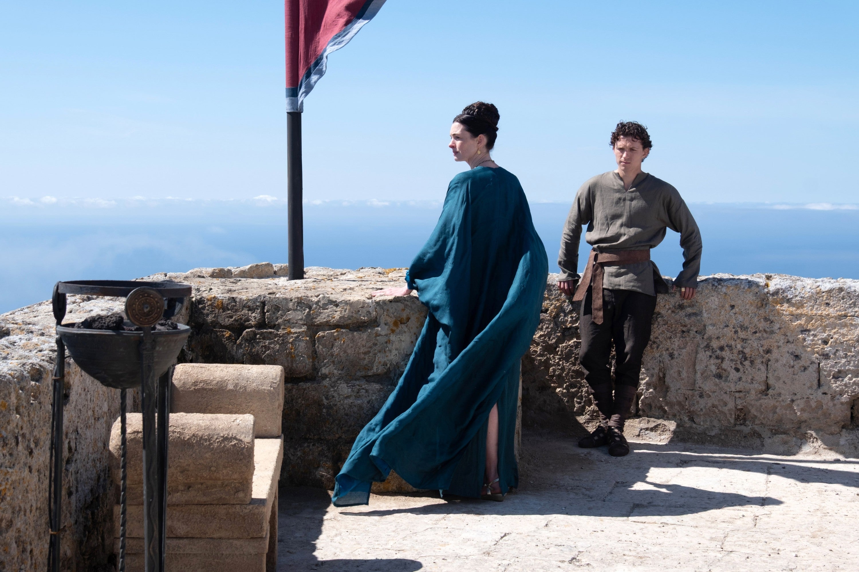 Two people in historical attire stand on a stone terrace by the sea, beneath a flag