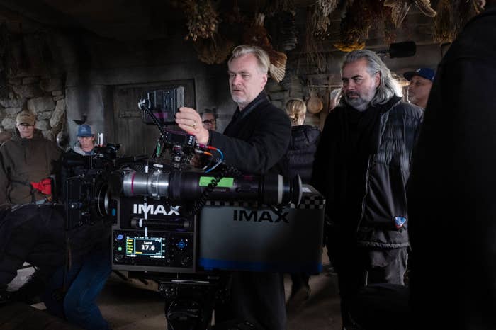A director and crew members work on a film set. The director looks at a monitor attached to an IMAX camera, surrounded by production equipment