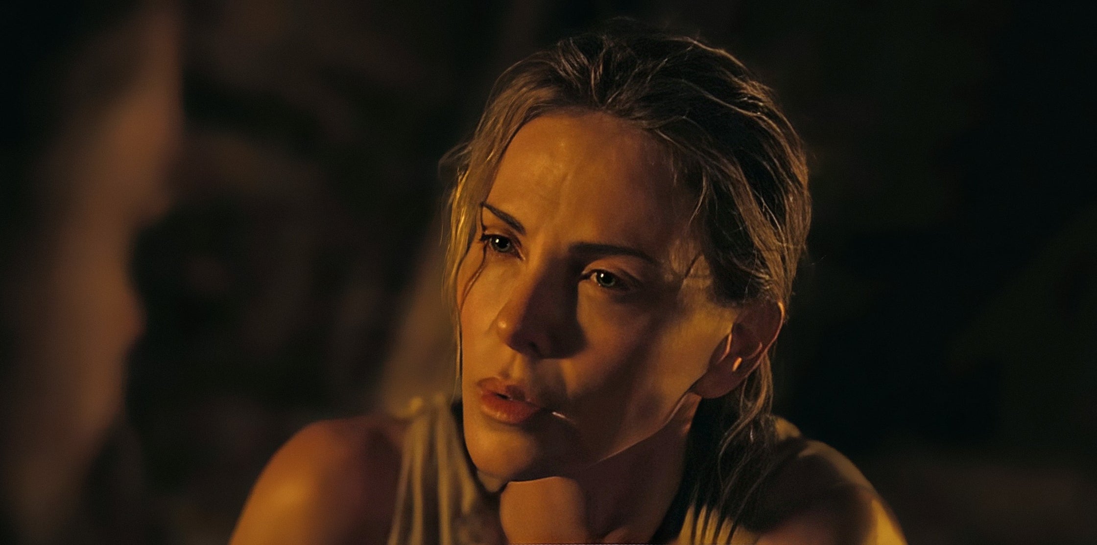 A person with a serious expression, wearing a sleeveless top, appears contemplative in a dimly lit setting