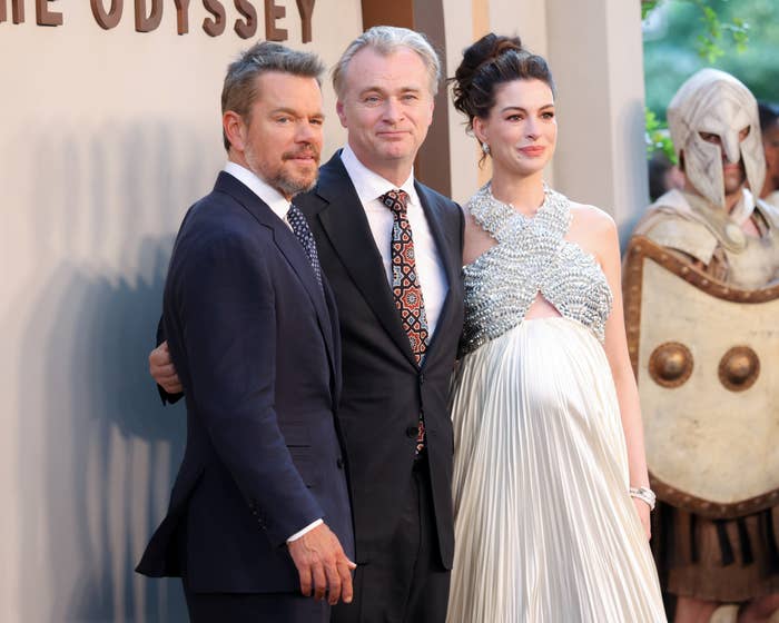 Three people pose at an event. One wears a patterned tie, and another is in an elegant sleeveless gown. A figure wearing armor is in the background
