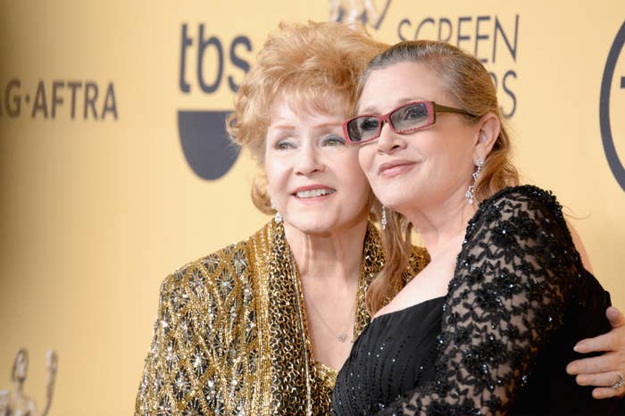 Two women pose together on a red carpet. One wears a sparkly jacket, the other a black dress with lace details. Both are smiling and embracing