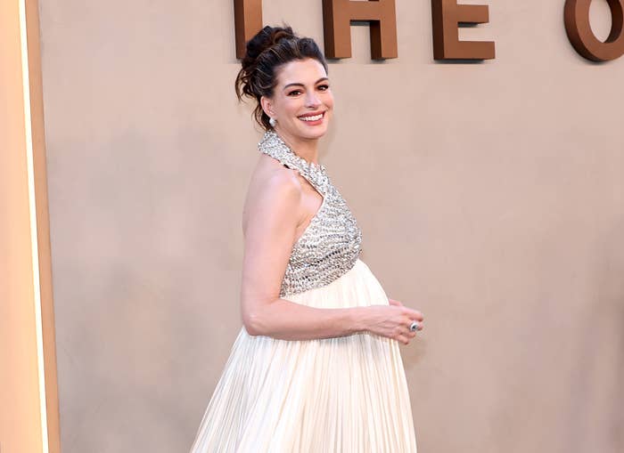 Person on a red carpet wearing a gown with a beaded bodice and pleated skirt, smiling and holding their baby bump