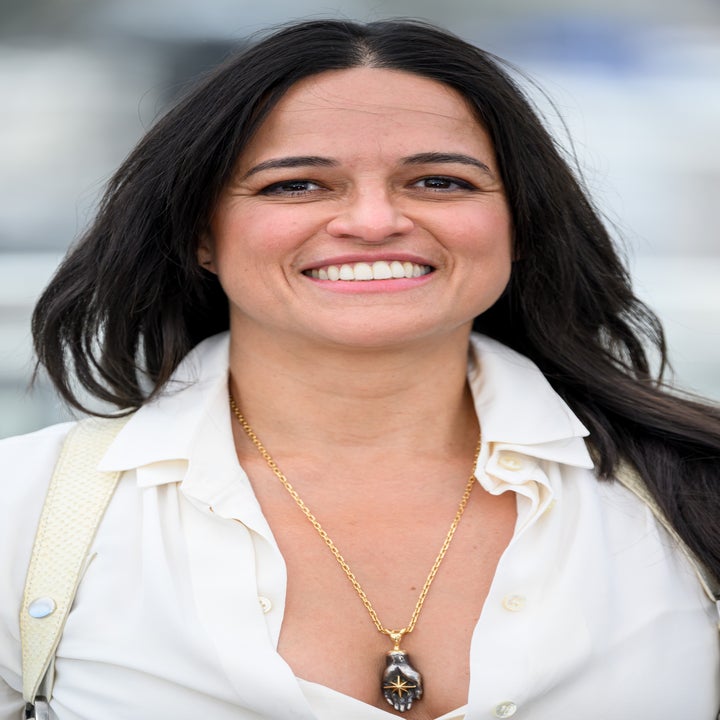 Person smiling, wearing a white blouse with a gold necklace, standing outdoors