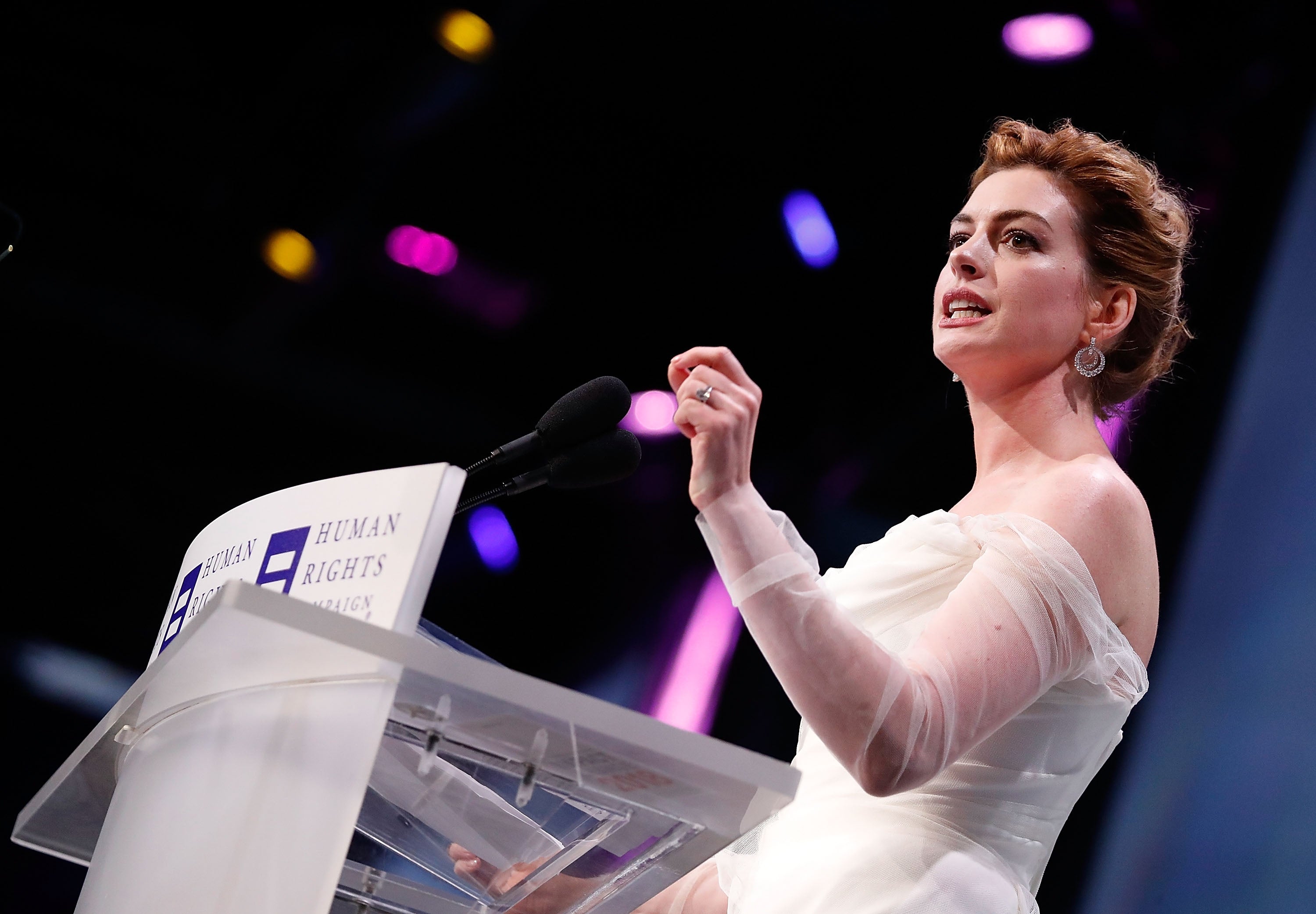 Person speaking at podium with "Human Rights" on it, elegantly dressed in an off-shoulder gown, addressing an audience
