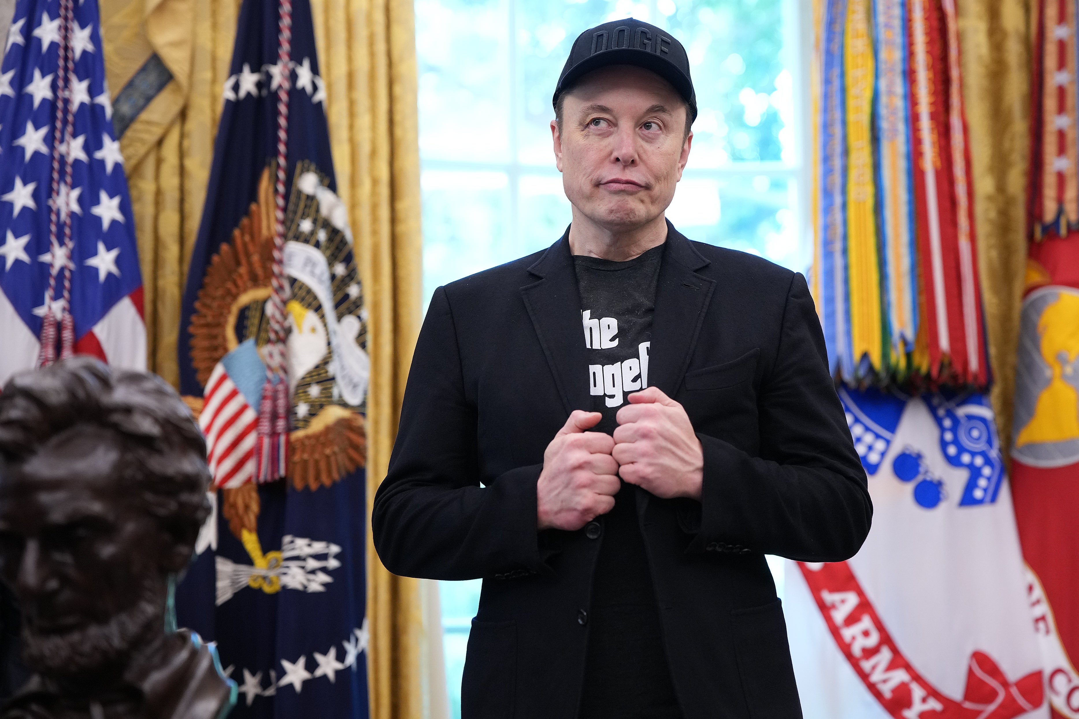 A person in a suit and cap stands confidently in an official room with military flags and a statue in the background