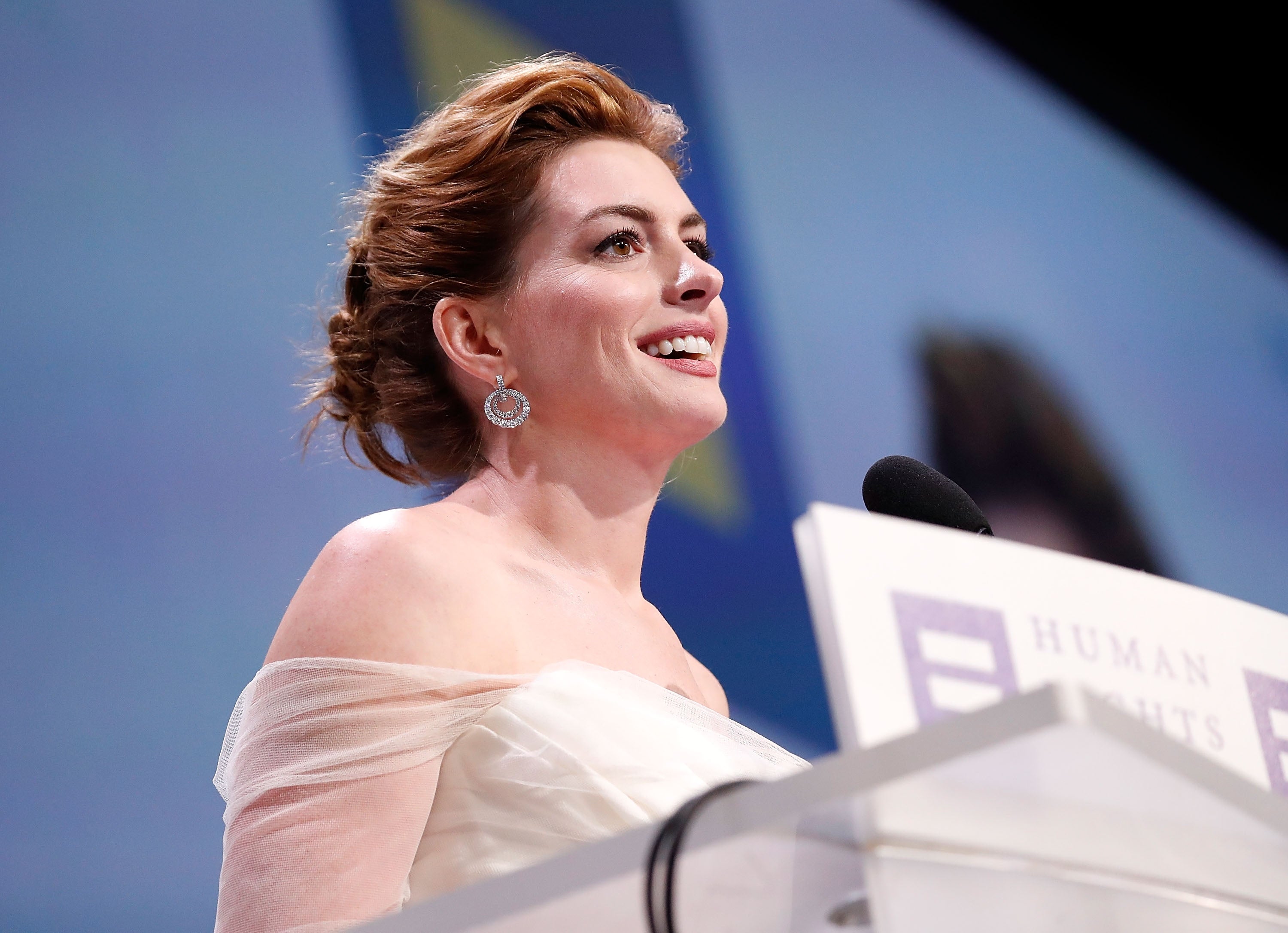 A person in a white gown delivers a speech at a podium, smiling, with Human Rights text visible on the podium