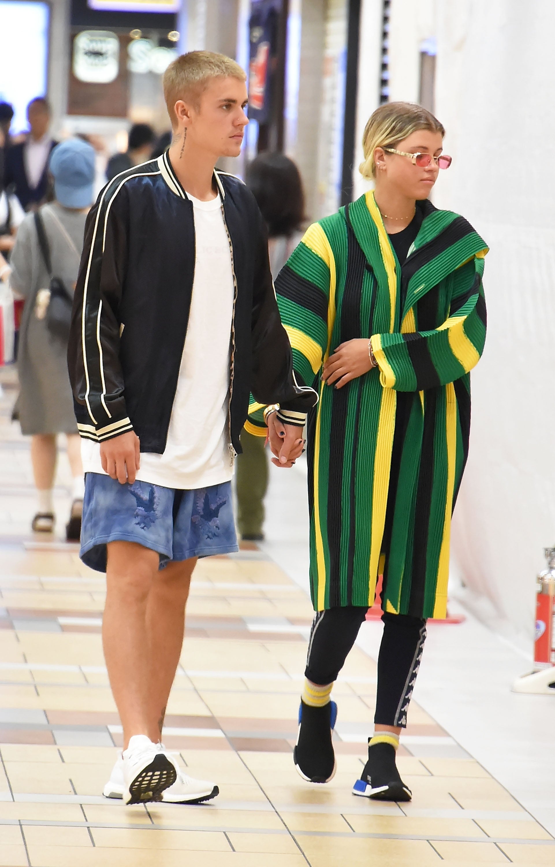 Two people walking and holding hands in a public area. One wears a sporty jacket and shorts, and the other wears a striped long coat with leggings