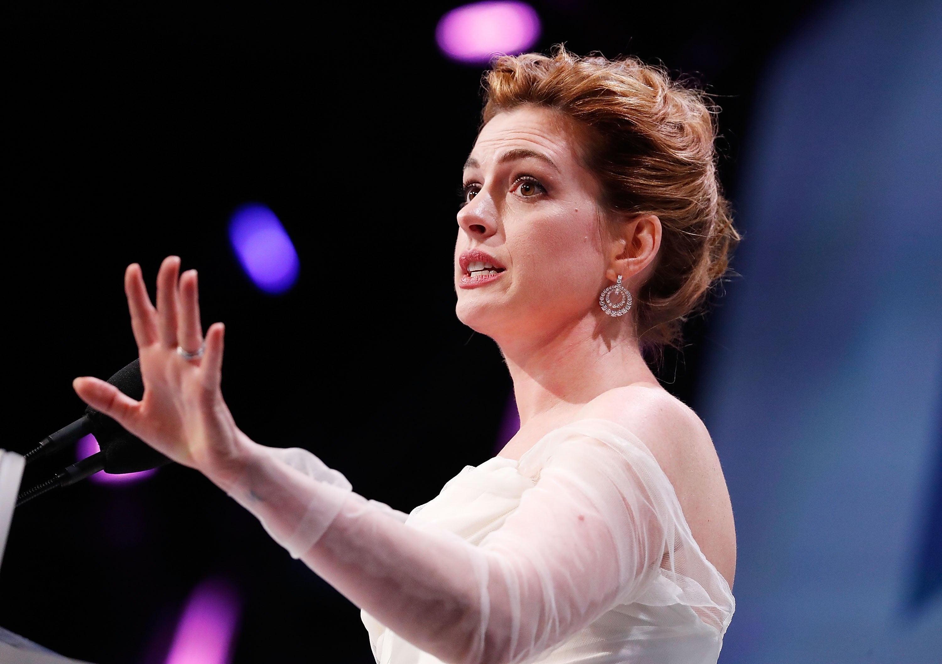 Person speaking at an event, wearing an elegant off-the-shoulder dress and statement earrings, with a dynamic gesture