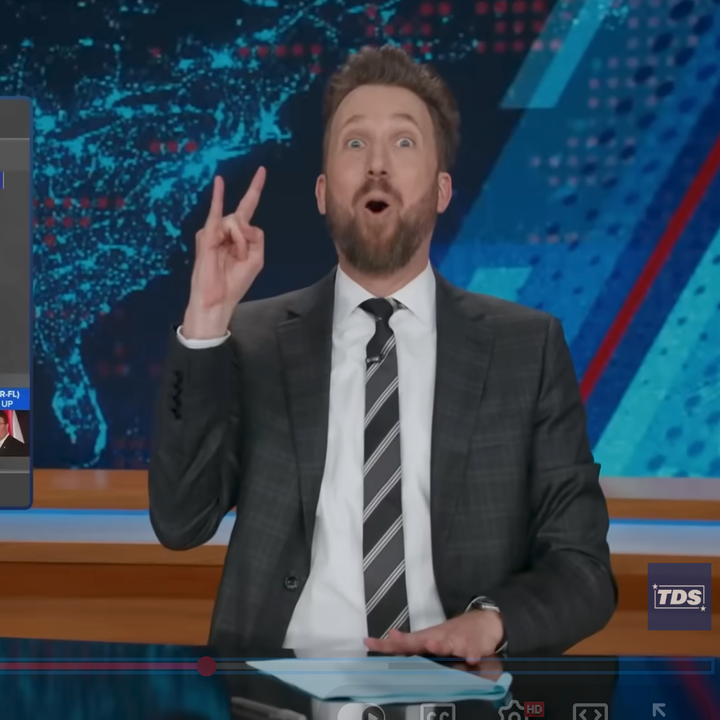 A person in a suit at a news desk makes a humorous gesture with their hand, expressing excitement or surprise