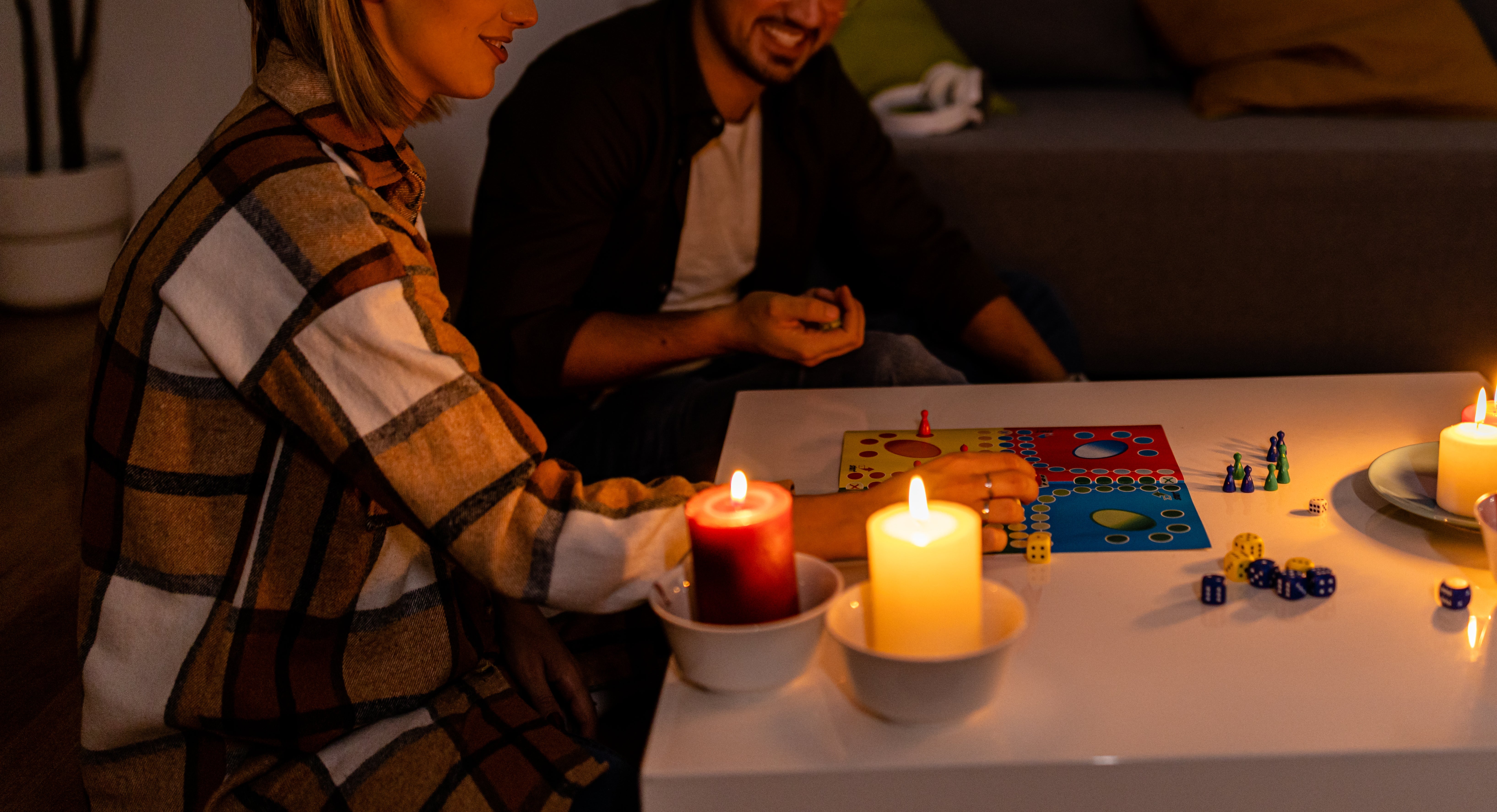 To personer spiller et brettspill ved et opplyst bord med stearinlys. Innstillingen er koselig, med en sofa og puter i bakgrunnen