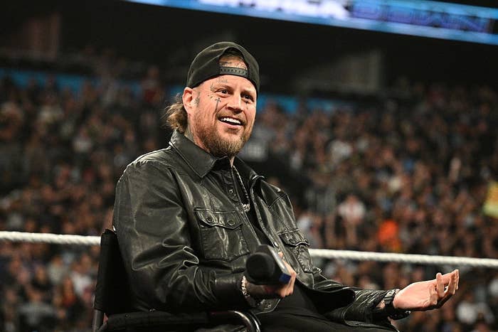 A person wearing a leather jacket and cap sits in a stadium, holding a microphone with a crowd in the background