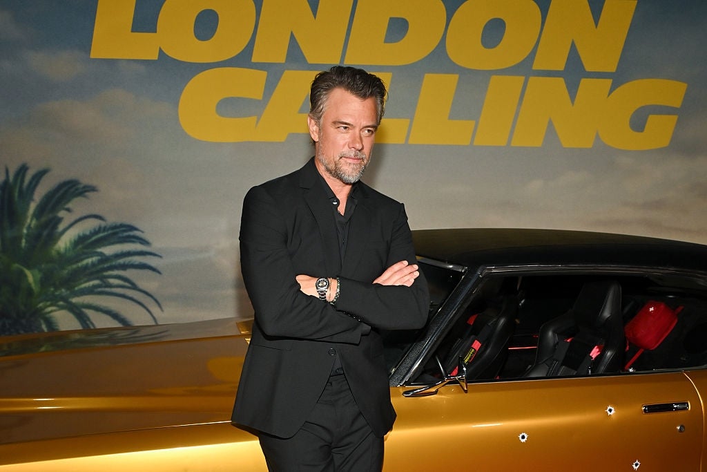 A man in a sleek black suit poses confidently next to a vintage car at a "London Calling" event backdrop