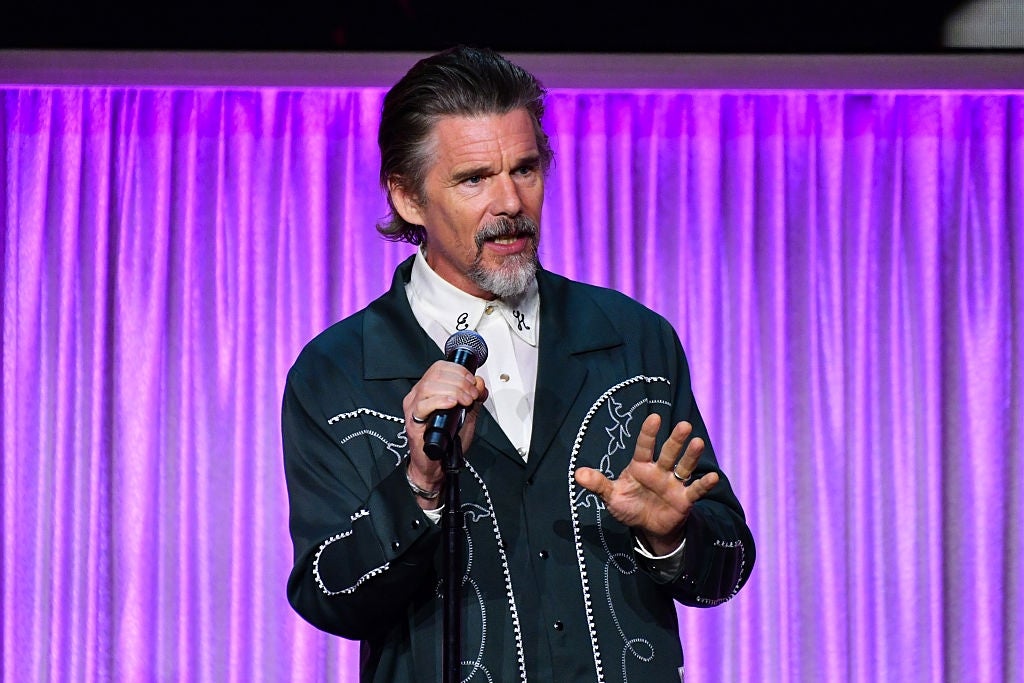 A celebrity in a western-style embroidered jacket speaks into a microphone on stage with a curtain backdrop
