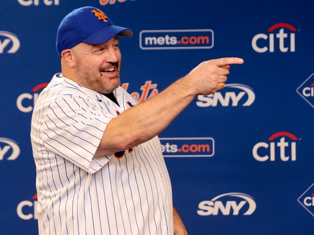 A person in a baseball cap and pinstripe jersey points and smiles at a Mets event backdrop
