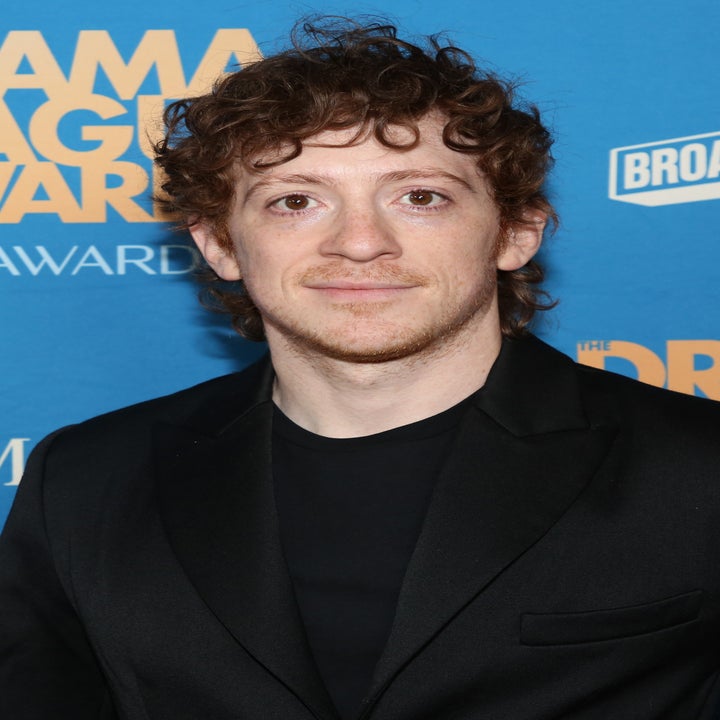 A person with curly hair in a black suit poses on the red carpet at an awards event