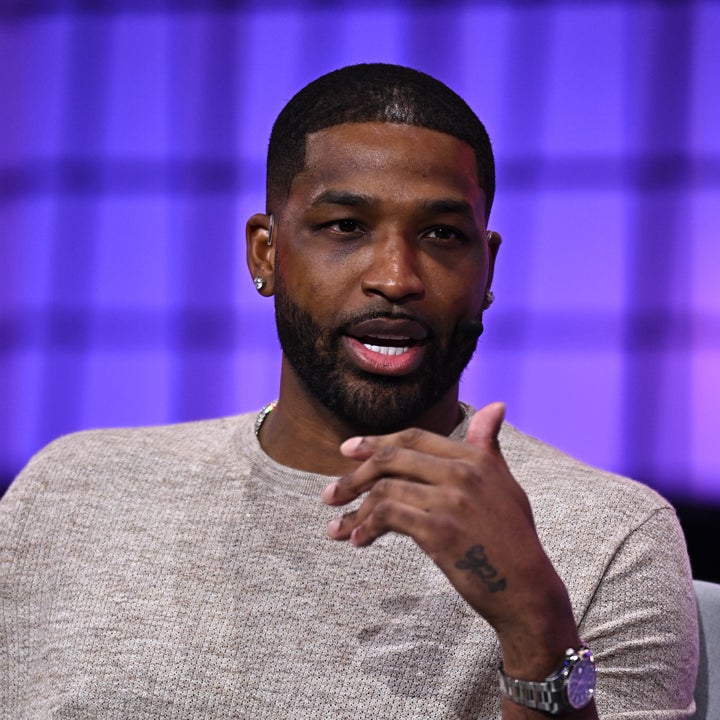 Person seated on a stage, wearing a casual short-sleeve shirt and pants, gesturing while speaking during an interview