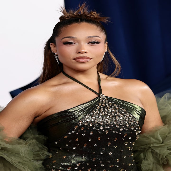 A person on the red carpet wears a shimmering, fitted gown with off-shoulder ruffled details and a sleek hairstyle, holding a small clutch