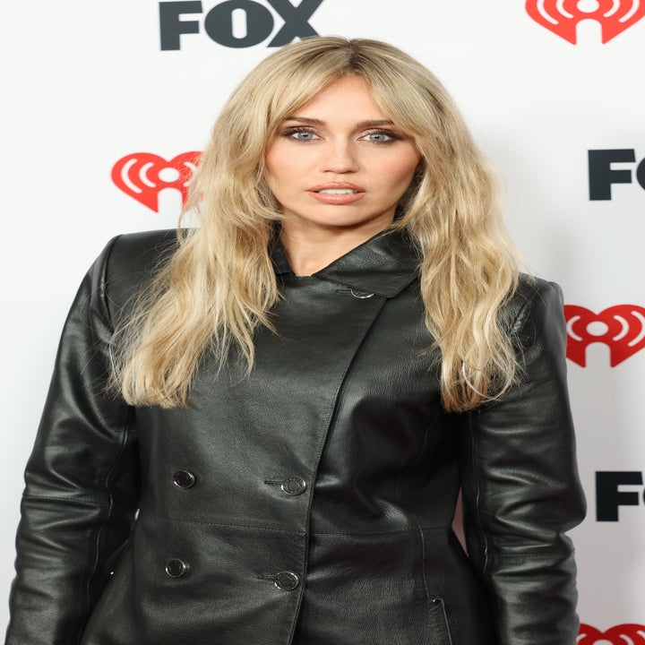 Person in a sleek leather coat at a celebrity event with logos in the background